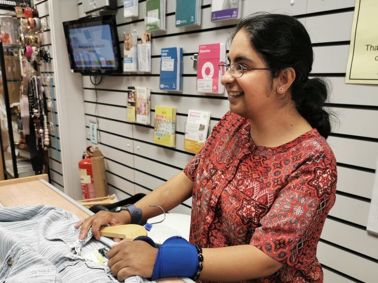 This shows our Bilston volunteer Heena stood at a till with a top in her hands.